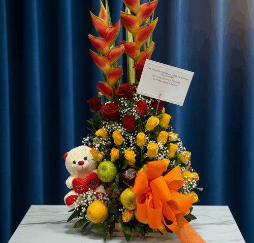 Arreglo floral vertical con rosas rojas y amarillas, heliconias naranjas, peluche blanco con corazón rojo, frutas variadas y gran lazo naranja sobre mesa de mármol