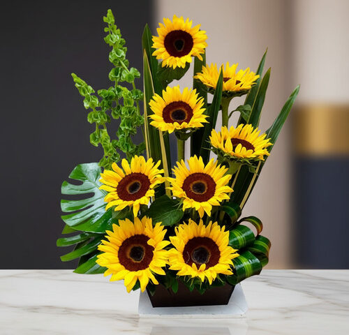Arreglo floral moderno con varios girasoles amarillos y follaje verde en base rectangular negra sobre mesa de mármol