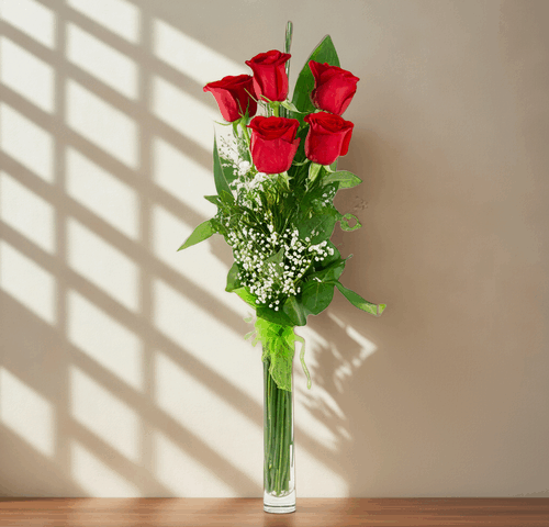 Seis rosas rojas con follaje verde y flores blancas pequeñas en un jarrón de vidrio alto y estrecho sobre una repisa de madera