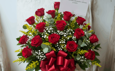 Canasta de mimbre con arreglo de rosas rojas, flores blancas pequeñas y follaje verde, decorada con lazo rojo y tarjeta sobre fondo claro