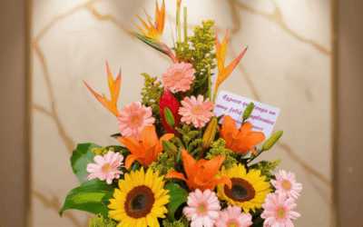Arreglo floral sobre base rectangular con girasoles amarillos, gerberas rosadas, lirios naranjas y hojas verdes, con una tarjeta de felicitación al fondo