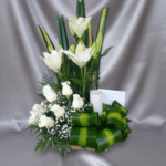 Arreglo floral vertical con lirios blancos altos, rosas blancas y follaje verde sobre tela gris