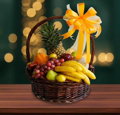 Canasta de mimbre sobre mesa de madera con frutas surtidas y gran lazo amarillo, fondo desenfocado con luces