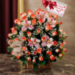 Canasta de mimbre sobre mesa de mármol con arreglo de rosas anaranjadas, lirios rosados y paniculata blanca, decorada con lazo beige y tarjeta colgante con mensaje de Happy Valentine’s Day