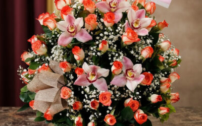 Canasta de mimbre sobre mesa de mármol con arreglo de rosas anaranjadas, lirios rosados y paniculata blanca, decorada con lazo beige y tarjeta colgante con mensaje de Happy Valentine’s Day