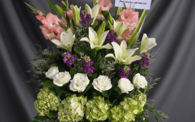 Arreglo floral grande con rosas blancas, lirios blancos, hortensias verdes, flores rosadas y moradas, con tarjeta de condolencias sobre fondo gris