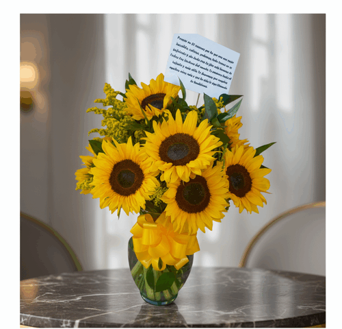 Arreglo de girasoles amarillos con lazo y tarjeta en un jarrón de cristal sobre una mesa de mármol en interior