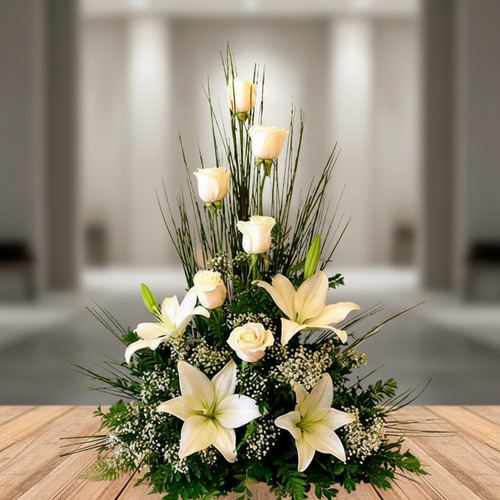 Centro de mesa con rosas y lirios blancos sobre una superficie de madera, con fondo interior desenfocado