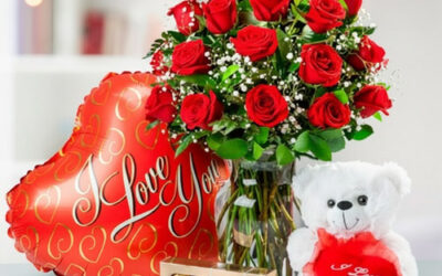 Ramo de rosas rojas en jarrón de cristal junto a globo rojo en forma de corazón con texto I Love You, oso de peluche blanco con corazón rojo y caja de chocolates Ferrero Rocher