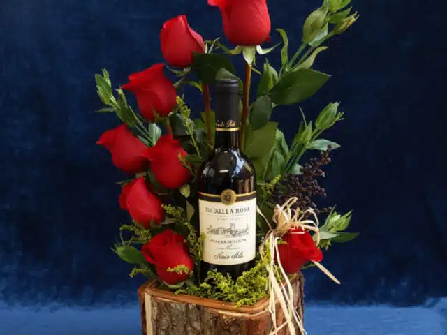 Arreglo floral VERITA con rosas rojas frescas y una botella de vino tinto, presentado en una base de madera rústica, ideal pa