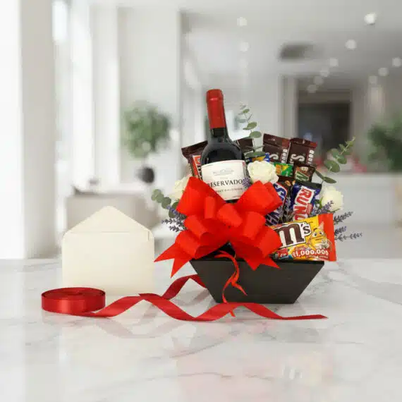 Caja Amadea con vino tinto, chocolates variados, rosas blancas y eucalipto, sobre mármol con tarjeta y cinta roja, lista para